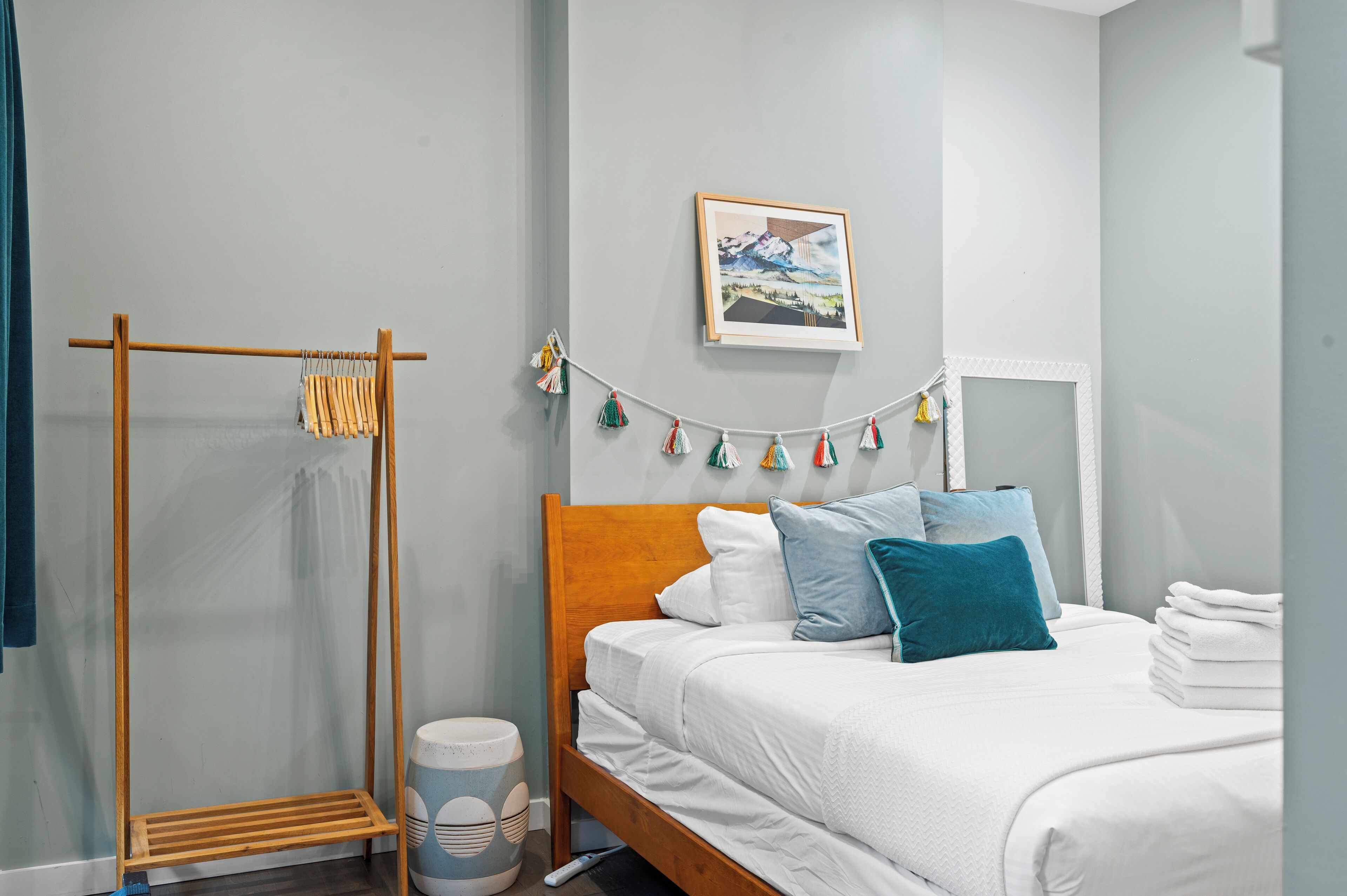 A neatly arranged bedroom with a bed, a wooden drying rack, a wall-mounted picture, and a string of colorful tassels.