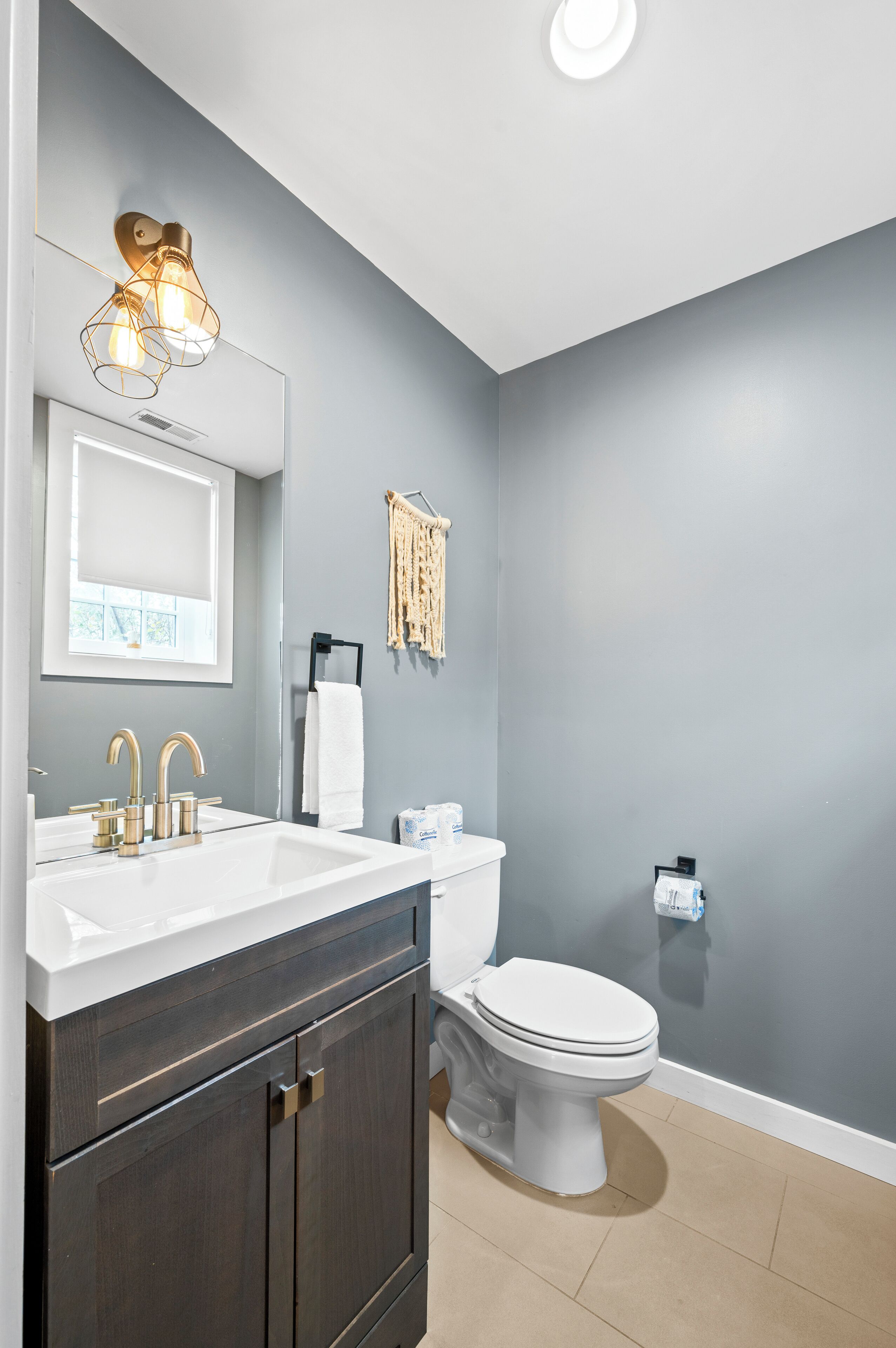 A modern bathroom with a dark gray wall, a white sink with gold fixtures, a white toilet, and a ceiling light fixture.