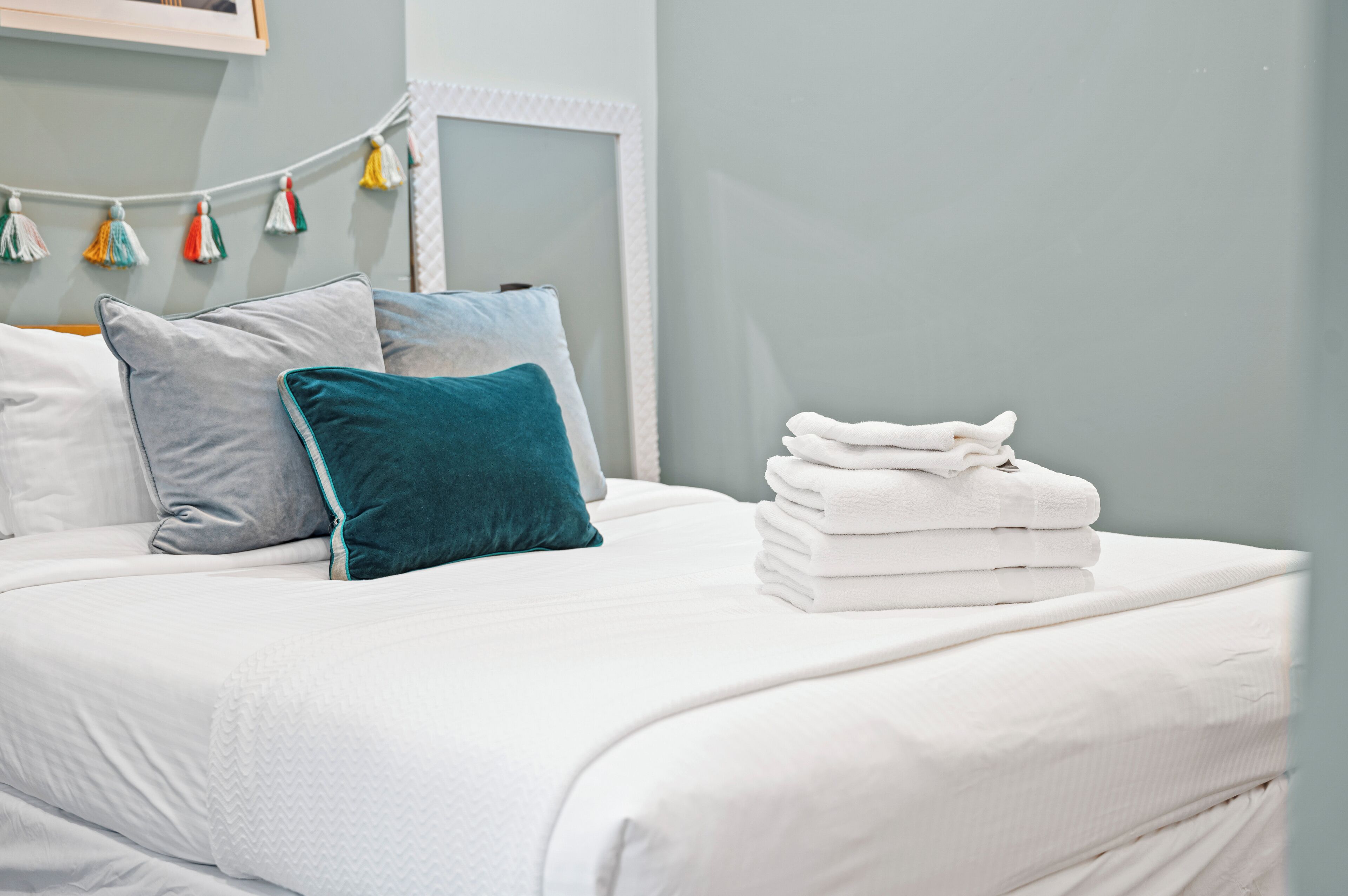 A neatly made bed with white linens and teal pillows, a stack of white towels, and a string of colorful tassels on the wall.