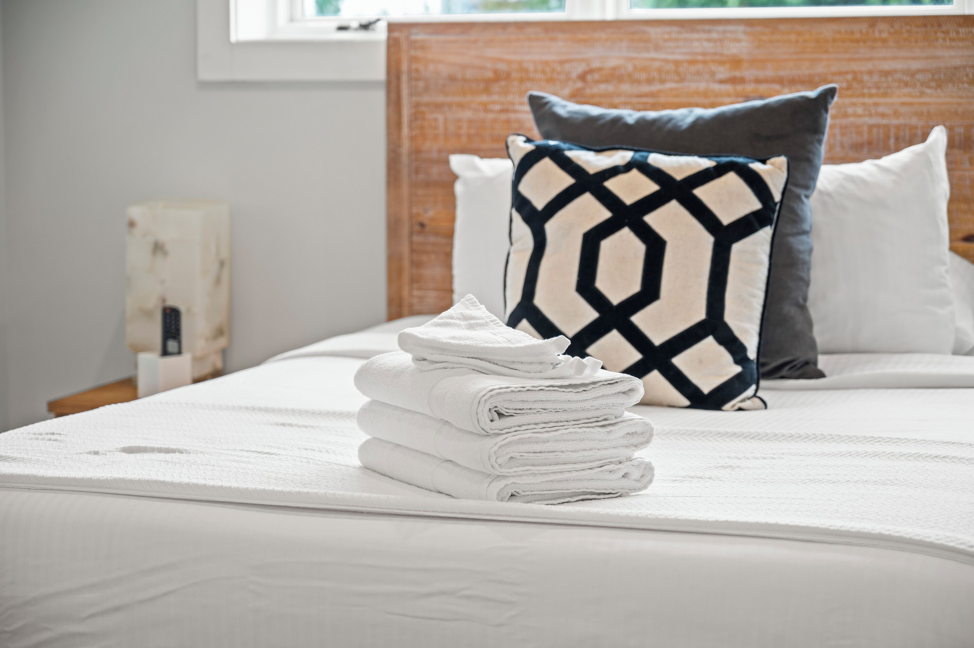 Un lit bien fait, avec un matelas blanc, une couverture blanche pliée et une pile de serviettes blanches. Il y a une tête de lit en bois, ornée d’un coussin à motifs géométriques et d’un coussin uni.