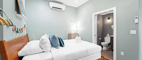 A neatly made bed with white linens and a wooden headboard, a bedside table with a lamp, and a wall-mounted air conditioner.