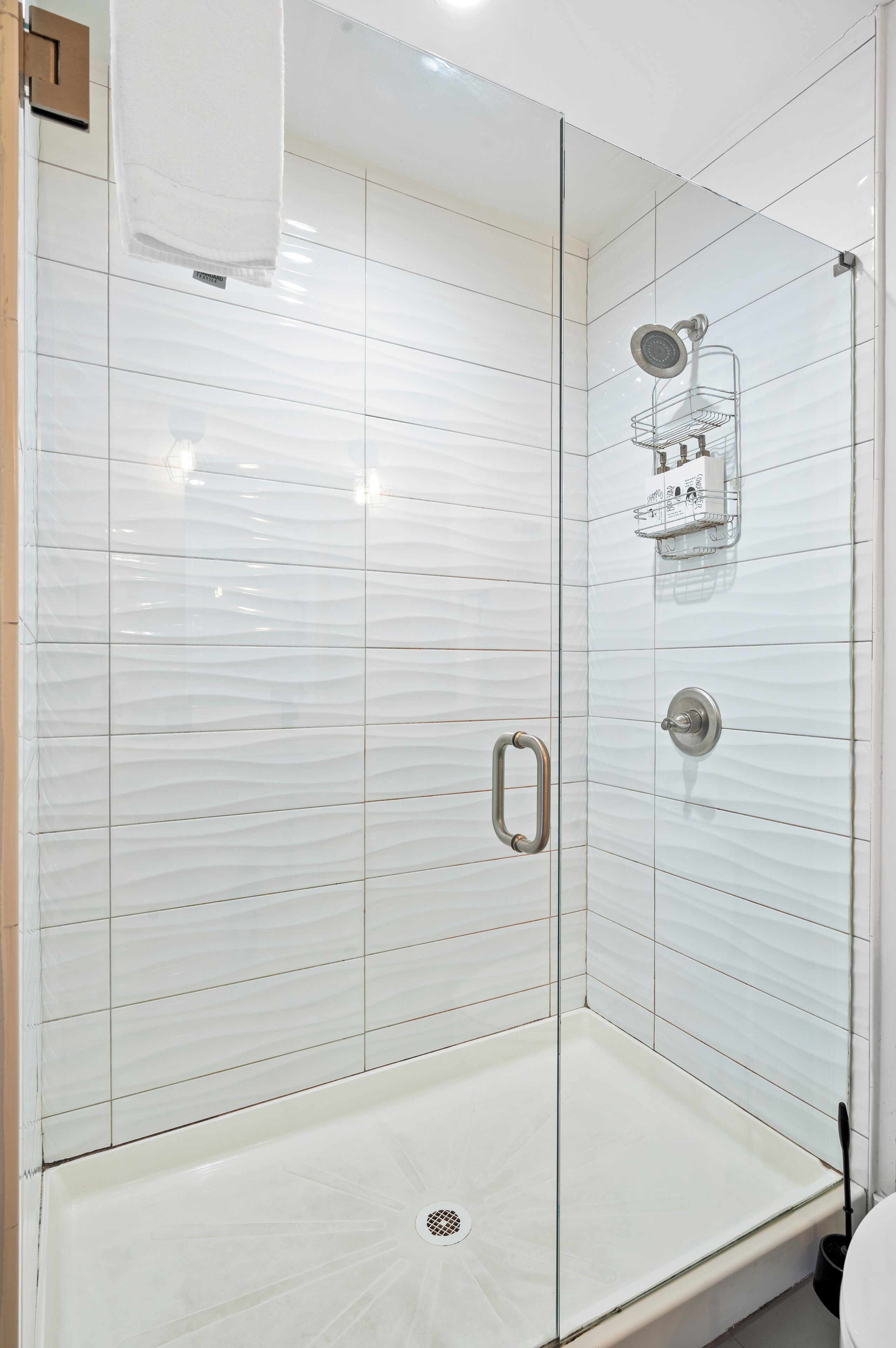 Ducha con cerramiento de vidrio, pared de azulejos blancos, una manija metálica y un cabezal de ducha.