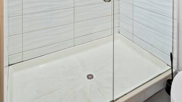 Ducha con cerramiento de vidrio, pared de azulejos blancos, una manija metálica y un cabezal de ducha.