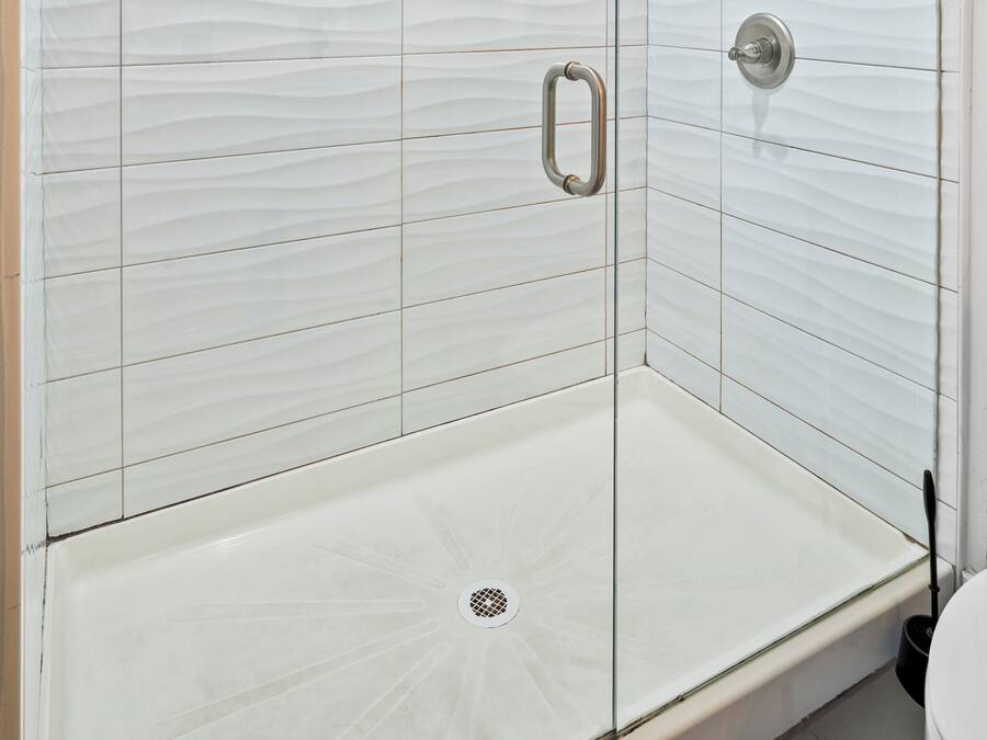 Ducha con cerramiento de vidrio, pared de azulejos blancos, una manija metálica y un cabezal de ducha.