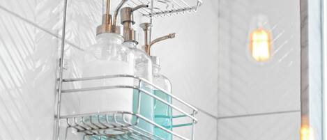 A bathroom with a shower head, a metal rack with bottles, and a light fixture on the wall.