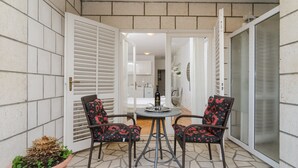 Um pátio externo com duas cadeiras de vime e uma mesa, um vaso de planta e vista para o interior da casa.