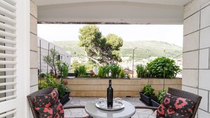 Um pátio externo com cadeiras de vime, uma mesa e uma garrafa de vinho. Há plantas em vasos e uma vista de uma colina ao fundo.