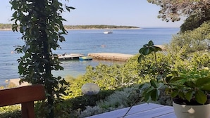 Une table en bois avec un pot de fleurs, un banc et une vue sur une mer calme où l’on aperçoit des bateaux.