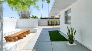 Eine moderne Außenpatio mit Betonboden, einer Holzbank, einer kleinen Grünfläche und einer hohen Pflanze.