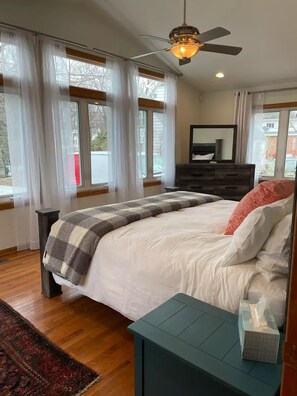 Un dormitorio con una cama, cómoda, mesita de noche, ventilador de techo y ventanas amplias con cortinas translúcidas.