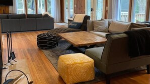 Un vaste salon doté d’un haut plafond, de poutres apparentes, d’un canapé d’angle gris, d’une table basse en bois et d’un pouf jaune.
