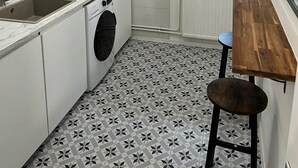 Une cuisine de taille réduite, équipée d’un lave-linge, d’un évier et d’une fenêtre avec des stores.