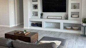 Ett modernt vardagsrum med en stor platt-TV, grå soffor, ett soffbord i trä och inbyggda hyllor med dekorativa föremål.