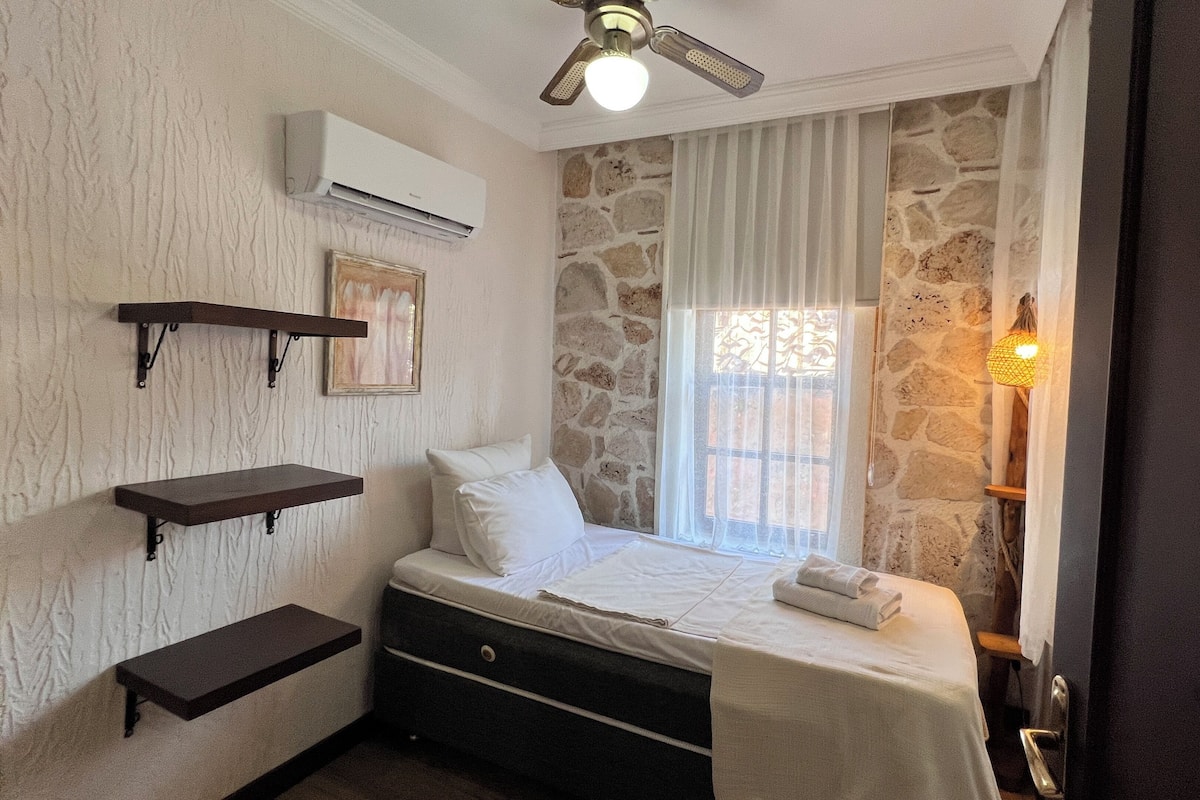A bedroom with a bed, a nightstand, a wall-mounted air conditioner, a ceiling fan, and a window with curtains.