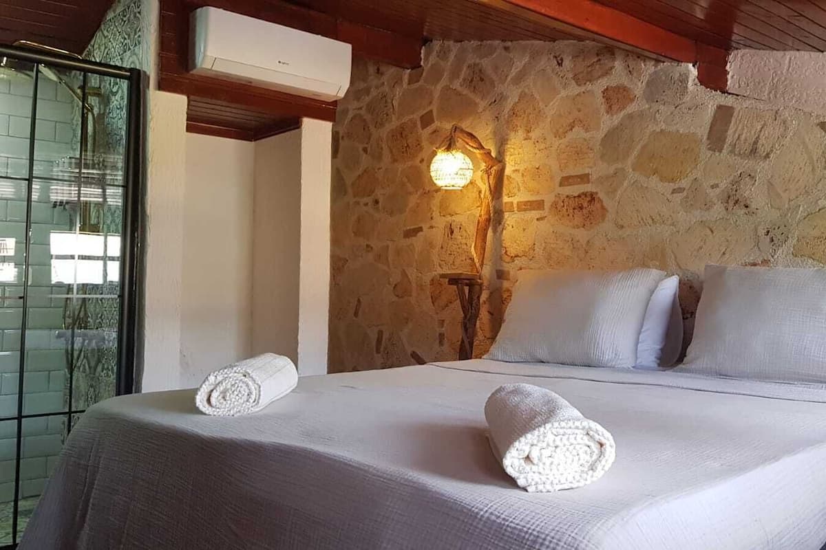 A bed with white linens and two rolled towels, a stone wall, and a wall-mounted light fixture.