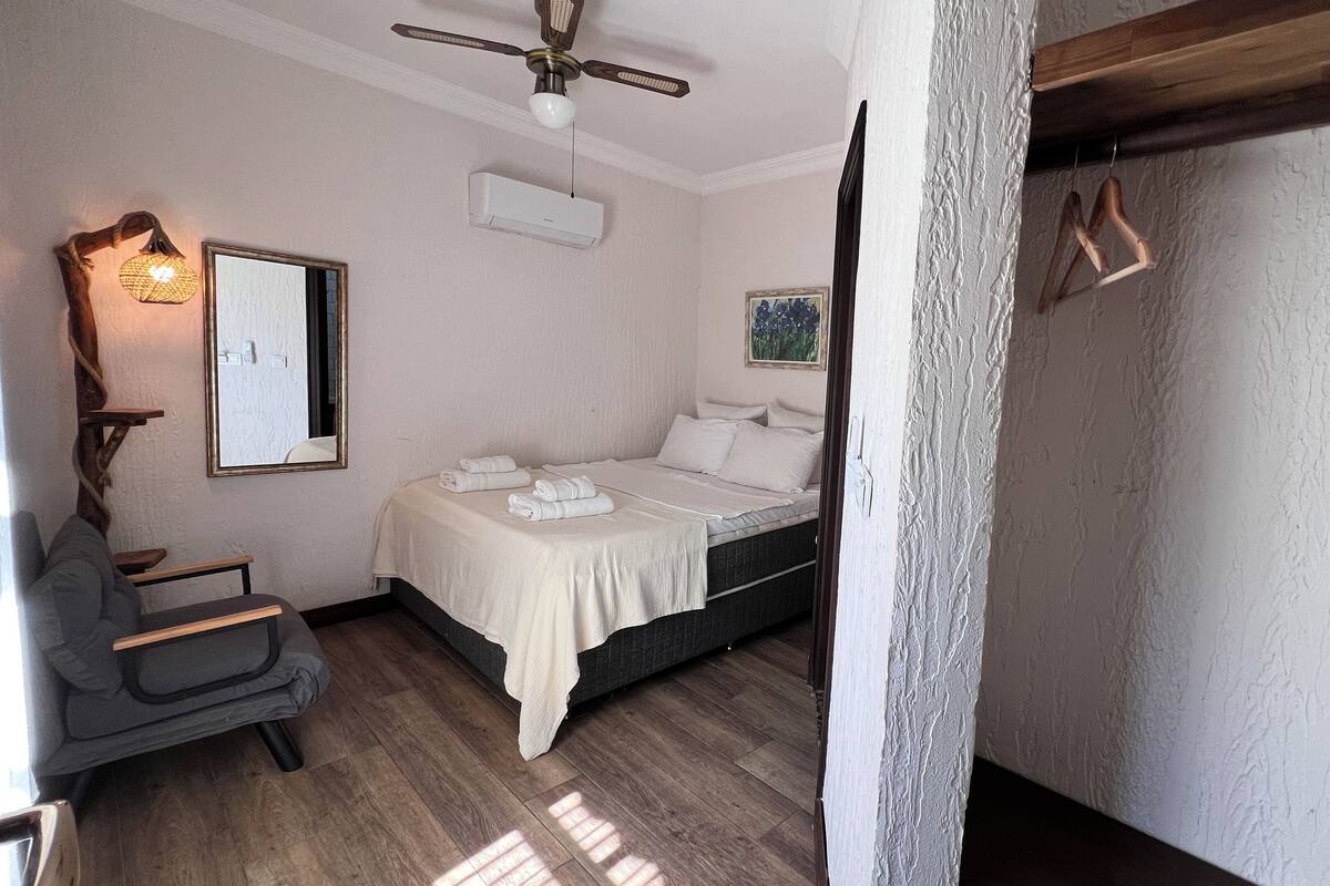 A bedroom with a bed, a chair, a ceiling fan, an air conditioning unit, and a mirror.
