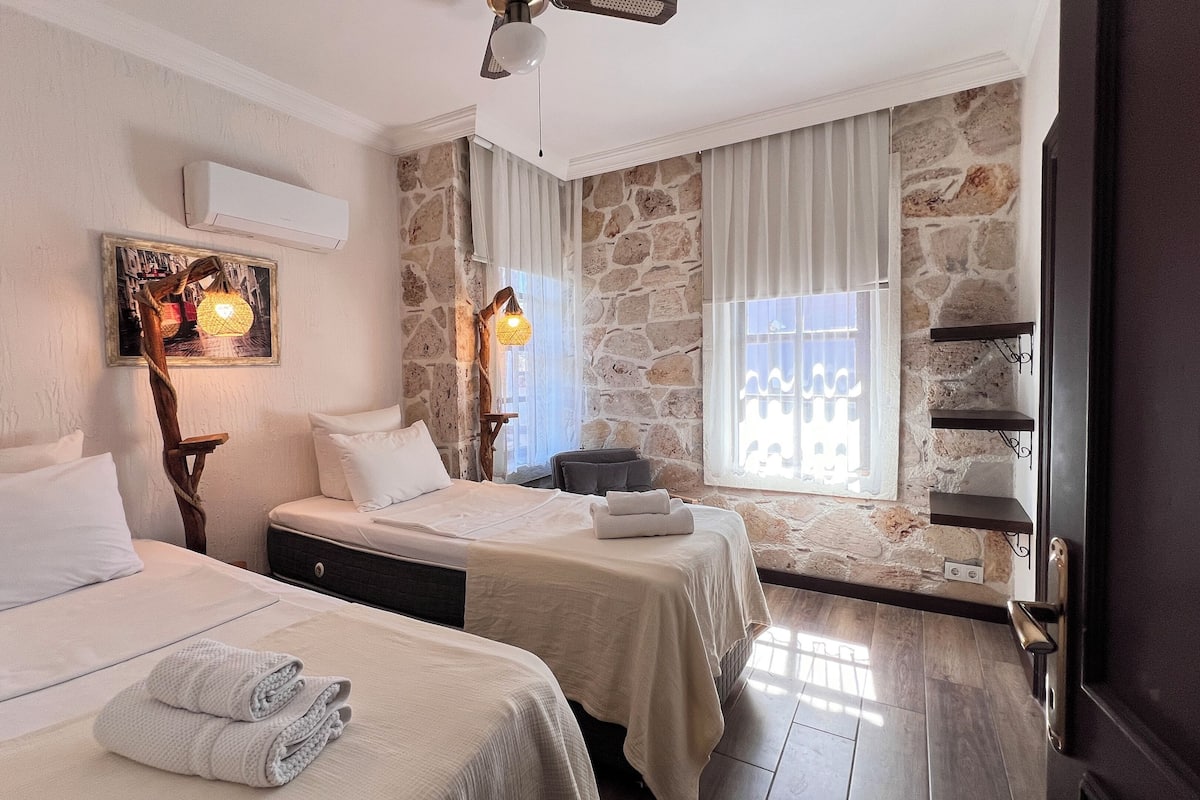 A hotel room with two beds, a stone wall, a large window with curtains, and a ceiling fan.
