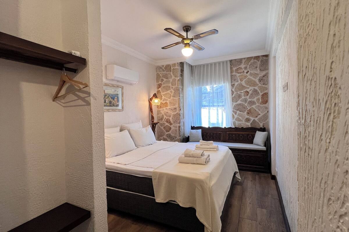 A bedroom with a bed, a bedside table, a wall-mounted air conditioner, a ceiling fan, a stone wall, a window with curtains, and a wooden bench.