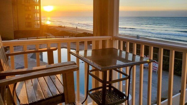 A balcony with a table and chairs, overlooking a beach at sunset. - Oceanfront views, Beachfront condo, The Driftwood! (north topsail beach)