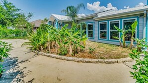 Une maison d’habitation à l’extérieur bleu, avec un jardin devant la maison où poussent des bananiers, et un ciel dégagé.