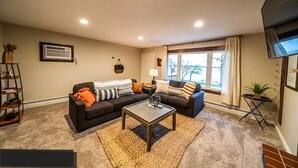 A living room with a sofa, coffee table, and a TV. There is a window with a view of trees and a balcony. - Lakefront Condo, 2 bedroom, with Dock & Private Patio! Walk to Restaurants! (Detroit Lakes)