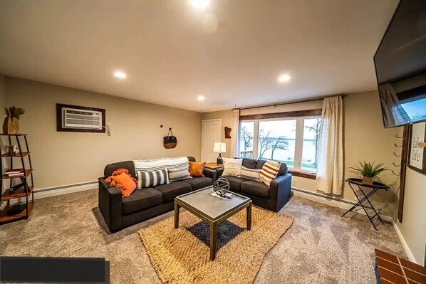 A living room with a sofa, coffee table, and a TV. There is a window with a view of trees and a balcony. - Lakefront Condo, 2 bedroom, with Dock & Private Patio! Walk to Restaurants! (Detroit Lakes)