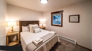 A neatly made bed with a wooden headboard, two bedside lamps, a window with a view, and a framed inspirational quote on the wall. - Lakefront Condo, 2 bedroom, with Dock & Private Patio! Walk to Restaurants! (Detroit Lakes)