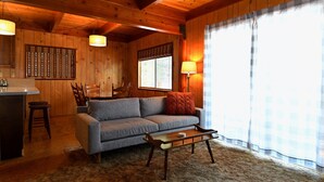A cozy cabin interior with a wooden dining table, a sofa, a coffee table, and a kitchen area with a sink and stools.