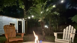 A backyard with a circular fire pit surrounded by rocks, two Adirondack chairs, and a wooden house with a door.