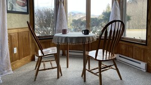 Een gezellige kamer met een houten tafel en twee stoelen, een ingelijst poster aan de muur en zicht op een besneeerd landschap door grote ramen.
