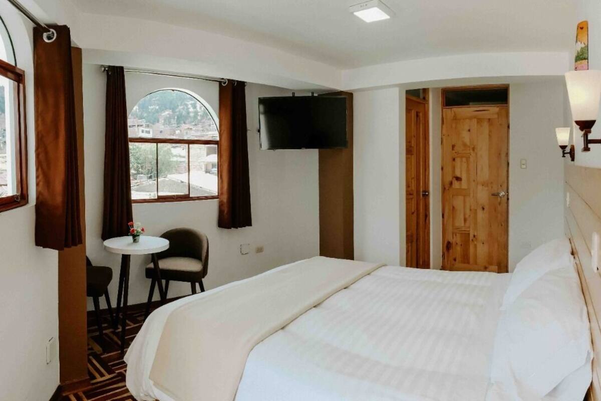Quarto de hotel com cama, televisão na parede, mesa pequena com duas cadeiras e janela com cortinas.