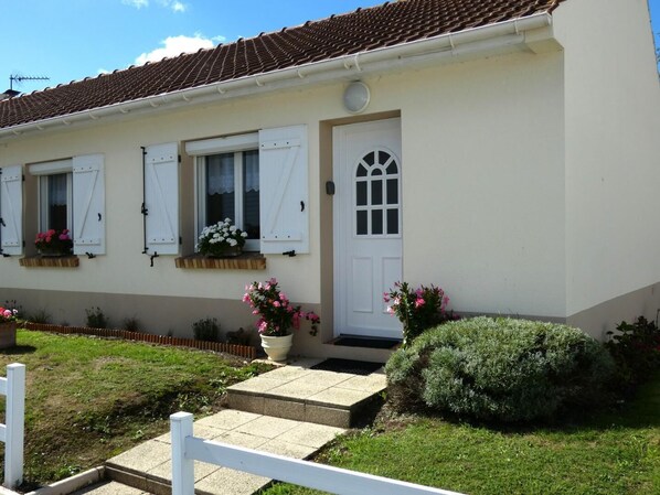 A two-story house with a tiled roof, white exterior walls, and a front door with a window above it. There are flower boxes on the windowsills and a small garden area with a white fence and a bush. - Gîte L'Escale - HESDIGNEUL LES BOULOGNE (HESDIGNEUL LES BOULOGNE)
