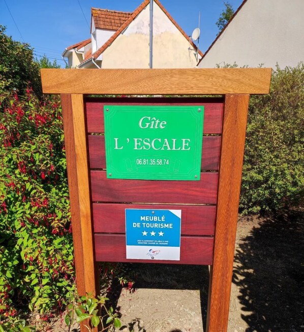 Et træskilt med en grøn markering med teksten "Gîte L'ESCALÉ" og en blå markering med teksten "MEUBLÉ DE TOURISME".