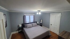 A bedroom with a bed, bedside tables, a window with curtains, and a door. - The Wesley - Historic 1890 Victorian, Second Floor (Albion)