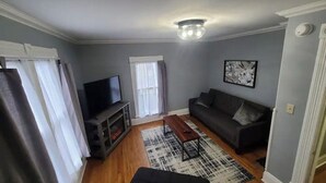 A living room with a sofa, a coffee table, a TV, and a fireplace. - The Wesley - Historic 1890 Victorian, Second Floor (Albion)