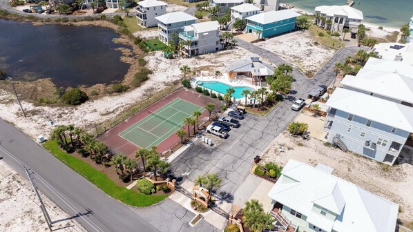 Vue aérienne d’un quartier résidentiel situé sur la côte, avec un court de tennis, une piscine et des propriétés donnant sur la plage.