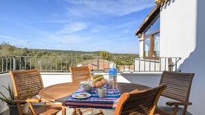 A round wooden table with four chairs, a colorful tablecloth, and a bottle of water on a balcony with a view of the countryside.