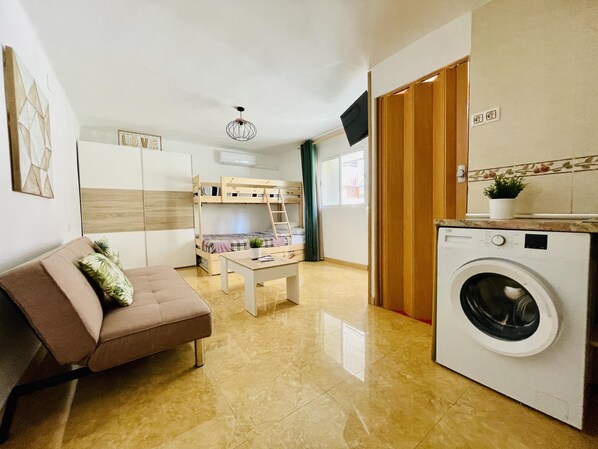 Un espai habitable reduït amb un sofà, lliteres, una rentadora i una televisió.