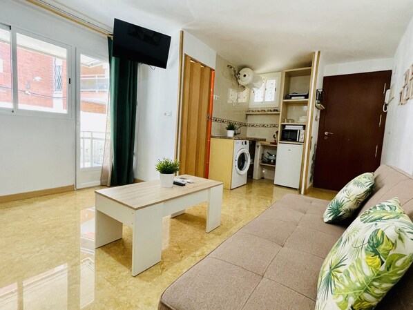 Un espacio habitable compacto con sofá, una mesita, lavadora, microondas y televisor montado en la pared.