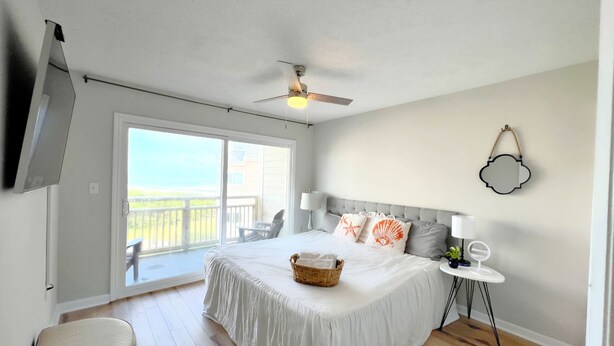 A bedroom with a bed, a ceiling fan, a bedside table with a lamp, a chair, and a view of the outdoors. - BEACHFRONT:3 BR, 2 Full BA, Renovated, Beach Access , BBQ, Pool, Sleeps 9 (Oak Island)