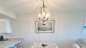A modern dining area with a wooden table, white chairs, a hanging chandelier, and a kitchen counter with stools. - BEACHFRONT:3 BR, 2 Full BA, Renovated, Beach Access , BBQ, Pool, Sleeps 9 (Oak Island)