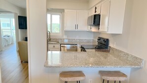 A modern kitchen with white cabinets, a granite countertop, a microwave, and a stainless steel stove. There are two wooden stools at the counter. - BEACHFRONT:3 BR, 2 Full BA, Renovated, Beach Access , BBQ, Pool, Sleeps 9 (Oak Island)