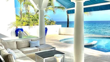 A sunlit outdoor lounge area with a pool, white columns, and a view of the ocean.