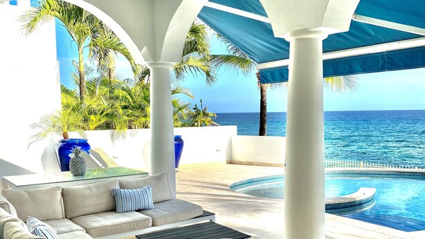 A sunlit outdoor lounge area with a pool, white columns, and a view of the ocean.