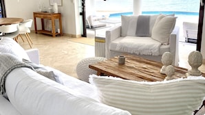 A modern living room with a large window overlooking a beach. There is a white sofa, a wooden coffee table, and a small side table with decorative items.