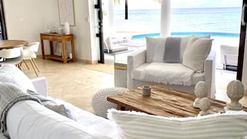 A modern living room with a large window overlooking a beach. There is a white sofa, a wooden coffee table, and a small side table with decorative items.