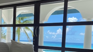 A room with a large window overlooking a beach, a white sofa, a wooden coffee table with a remote control, and a pair of sneakers.