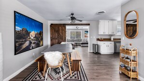 A modern kitchen with white cabinets, a dining area with a wooden table and bench, and a large painting on the wall. - Lucky Moon Retreat | JTree Oasis w/Hot Tub & Pool (Yucca Valley)