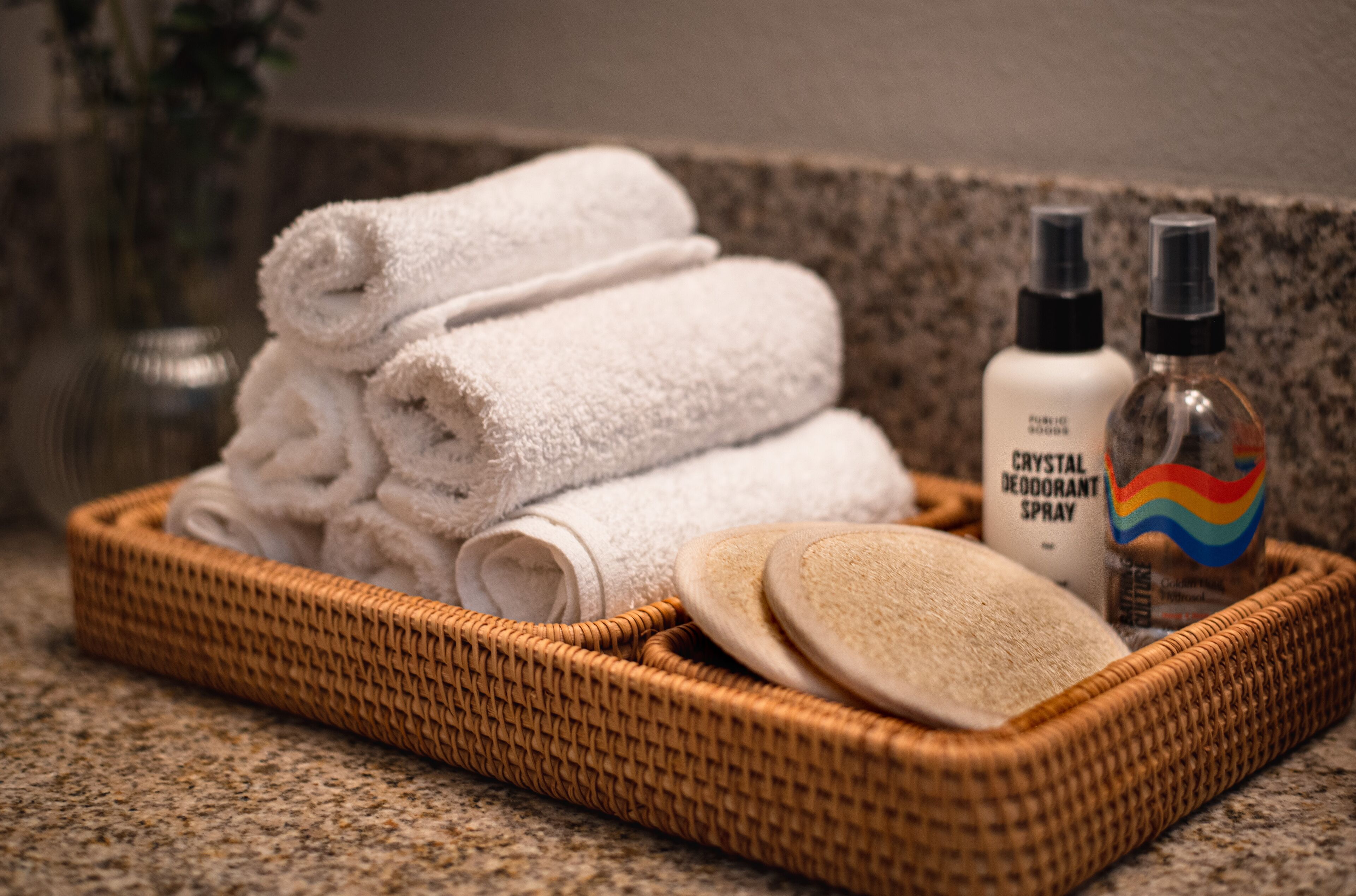 A wicker tray with neatly folded white towels, a bottle of crystal deodorant spray, and two wooden bath mats on a granite countertop.