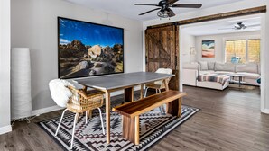 A dining area with a wooden table, chairs, and a bench. There is a large painting on the wall, a ceiling fan, and a view into another room with a couch. - Lucky Moon Retreat | JTree Oasis w/Hot Tub & Pool (Yucca Valley)